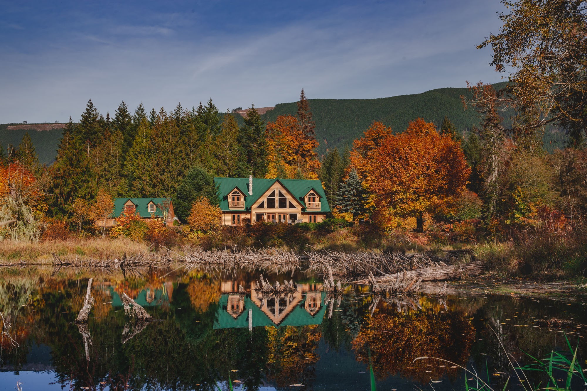 Een blokhutlodge met groene daken weerspiegelt in het stille water van de Cowichan River, omgeven door levendige herfstkleuren in tinten oranje en goud, met beboste heuvels op de achtergrond onder een blauwe hemel bij Lake Cowichan, Vancouver Island, Brits-Columbia, Canada.