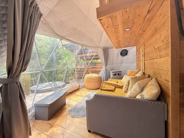 Gemütliches Inneres einer geodätischen Kuppel-Glamping-Unterkunft mit Sofa, goldenen Kissen, einem Pouf und einem kleinen Holzofen, mit einem großen dreieckigen Fenster mit Blick auf eine bewaldete Terrasse mit Whirlpool im Saguenay-Fjord-Nationalpark, Quebec, Kanada.