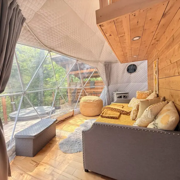 Cosy interior of a geodesic dome glamping accommodation with a sofa, golden cushions, a pouf, and a small wood stove, featuring a large triangular window overlooking a forested deck with a hot tub at Saguenay Fjord National Park, Quebec, Canada.