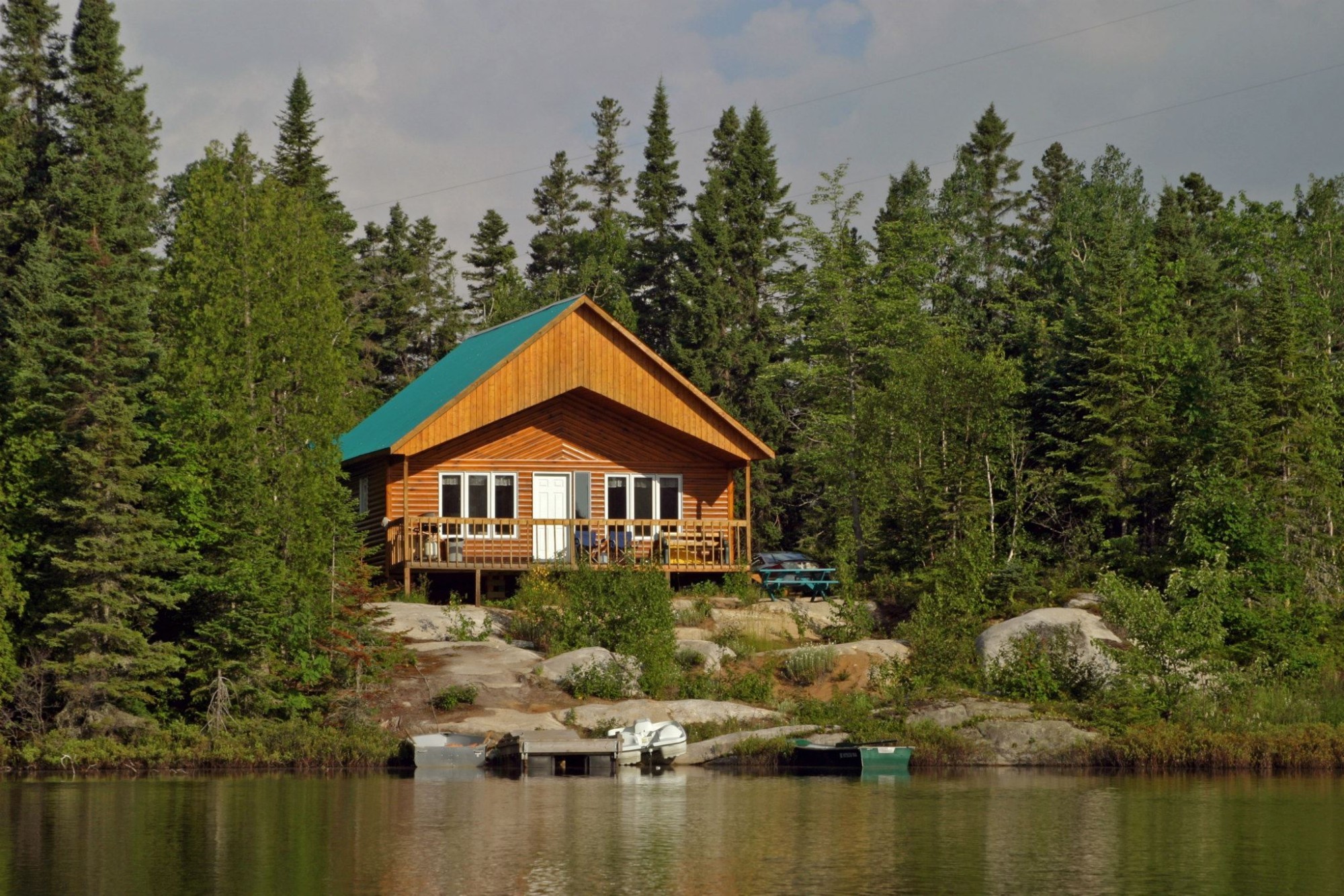 Een houten hut met een blauwgroen dak en omringend terras staat op een rotsachtige oever, omgeven door dicht dennenbos, met kleine boten aangemeerd aan een steiger op de voorgrond bij Tadoussac, Quebec, Canada.