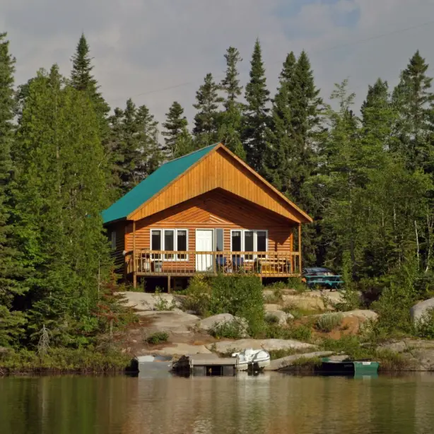 Een houten hut met een blauwgroen dak en omringend terras staat op een rotsachtige oever, omgeven door dicht dennenbos, met kleine boten aangemeerd aan een steiger op de voorgrond bij Tadoussac, Quebec, Canada.