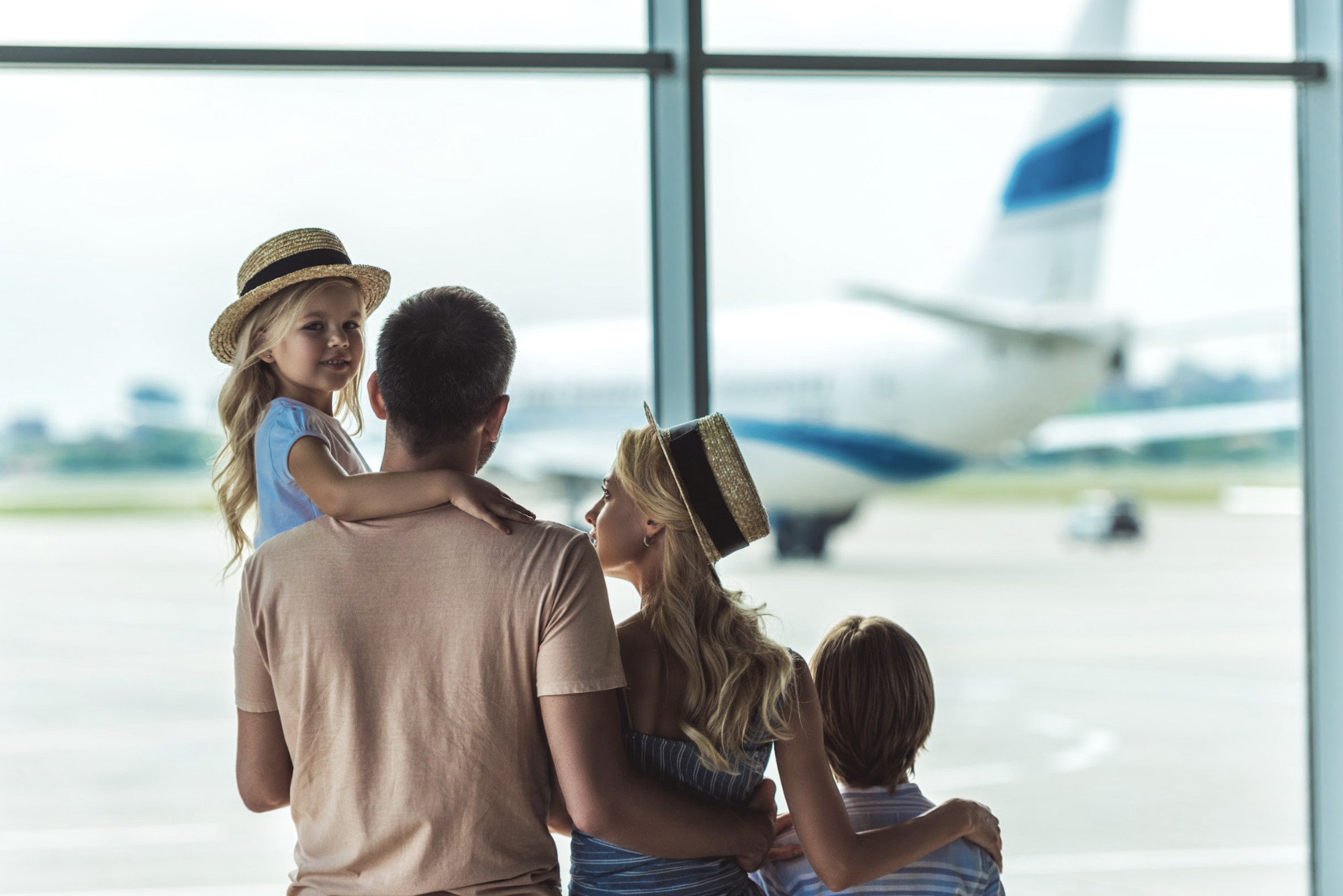 Een gezin met twee jonge kinderen dat vanuit het raam van een luchthaventerminal naar een vliegtuig op het platform kijkt, dat klaarstaat om op vakantie te vertrekken