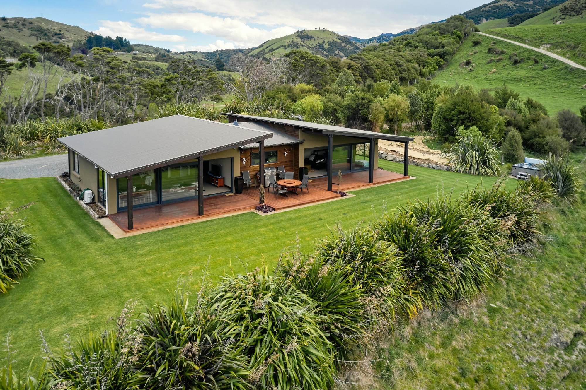Modernes Ferienhaus mit breiter überdachter Terrasse und Außenessbereich, auf einem gepflegten Rasen umgeben von einheimischen Pflanzen und sanften grünen Hügeln in der Nähe von Kaikoura, Neuseeland