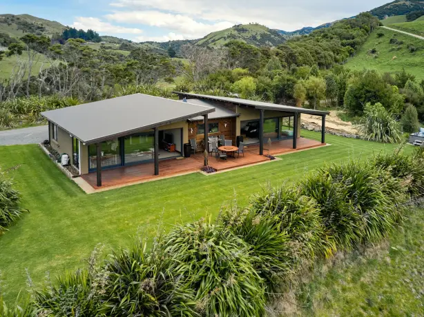 Modernes Ferienhaus mit breiter überdachter Terrasse und Außenessbereich, auf einem gepflegten Rasen umgeben von einheimischen Pflanzen und sanften grünen Hügeln in der Nähe von Kaikoura, Neuseeland