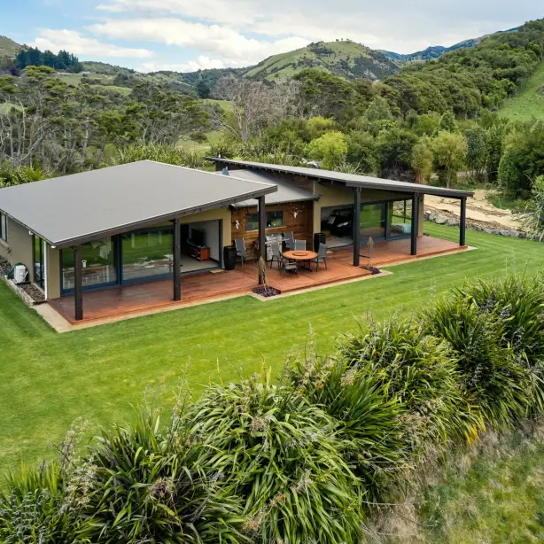 Modern holiday house with a wide covered deck and outdoor dining area, set on a manicured lawn surrounded by native plants and rolling green hills near Kaikoura, New Zealand