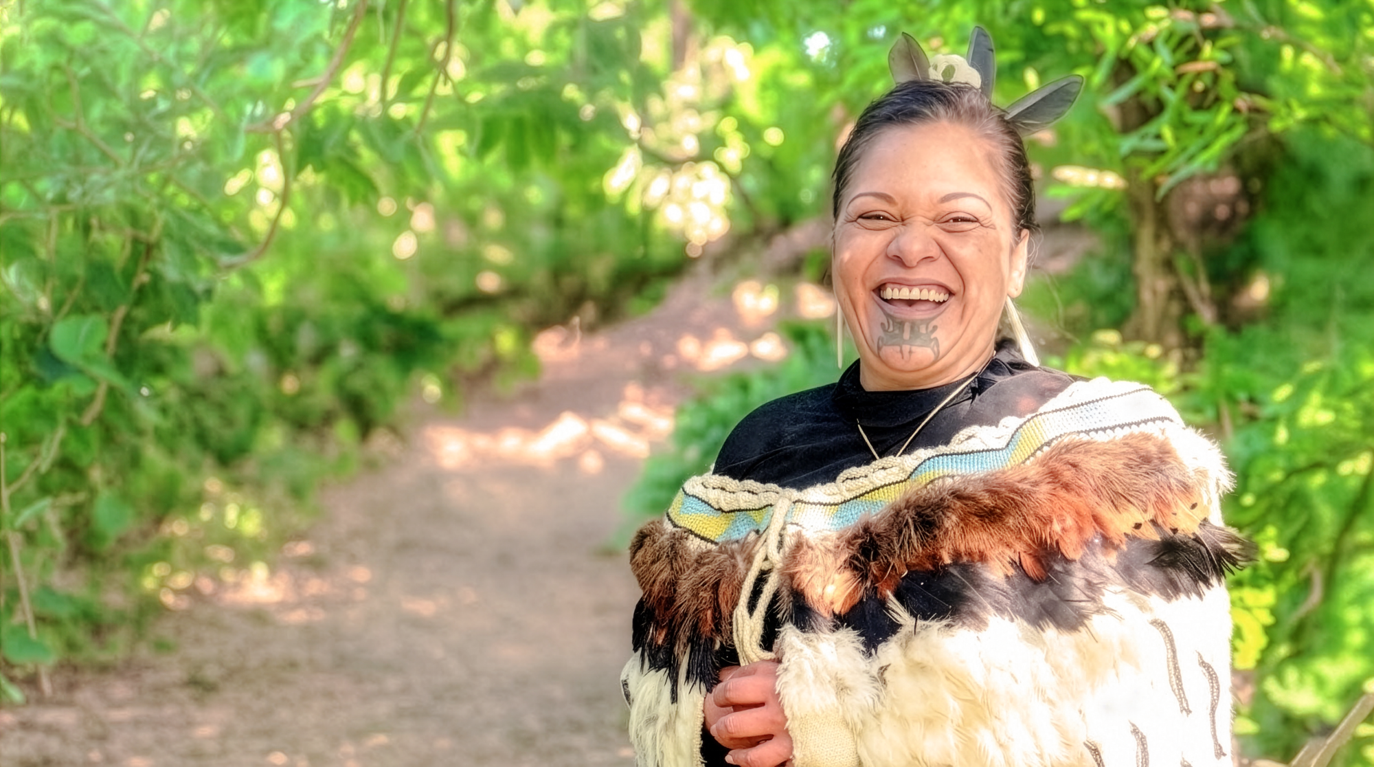 Smiling Māori woman Maria wearing a traditional feather cloak and tā moko facial tattoo, welcoming visitors as an indigenous local host in New Zealand