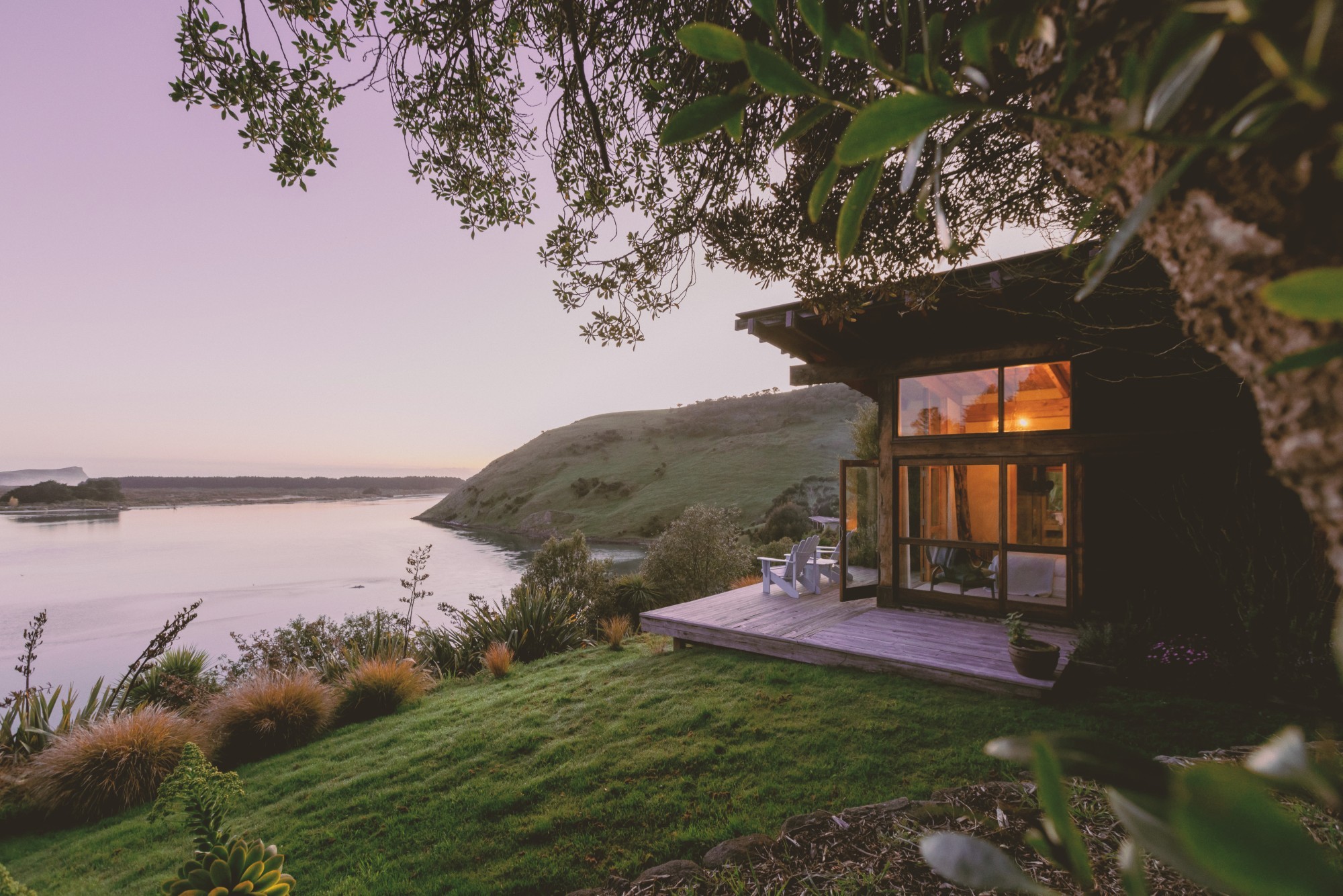 Ein abgeschiedenes Ferienhäuschen aus Holz mit einer Holzterrasse und zwei Stühlen, das an einem Hang thront und den Blick auf das ruhige Wasser der Otago-Halbinsel in Neuseeland bietet – in der Abenddämmerung, mit warm leuchtenden Innenräumen