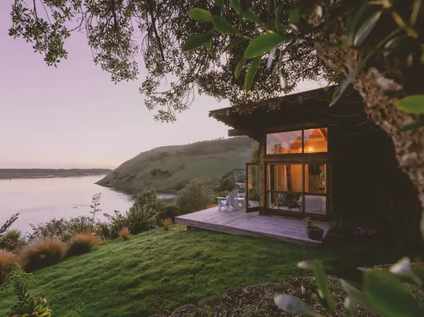 Ein abgeschiedenes Ferienhäuschen aus Holz mit einer Holzterrasse und zwei Stühlen, das an einem Hang thront und den Blick auf das ruhige Wasser der Otago-Halbinsel in Neuseeland bietet – in der Abenddämmerung, mit warm leuchtenden Innenräumen