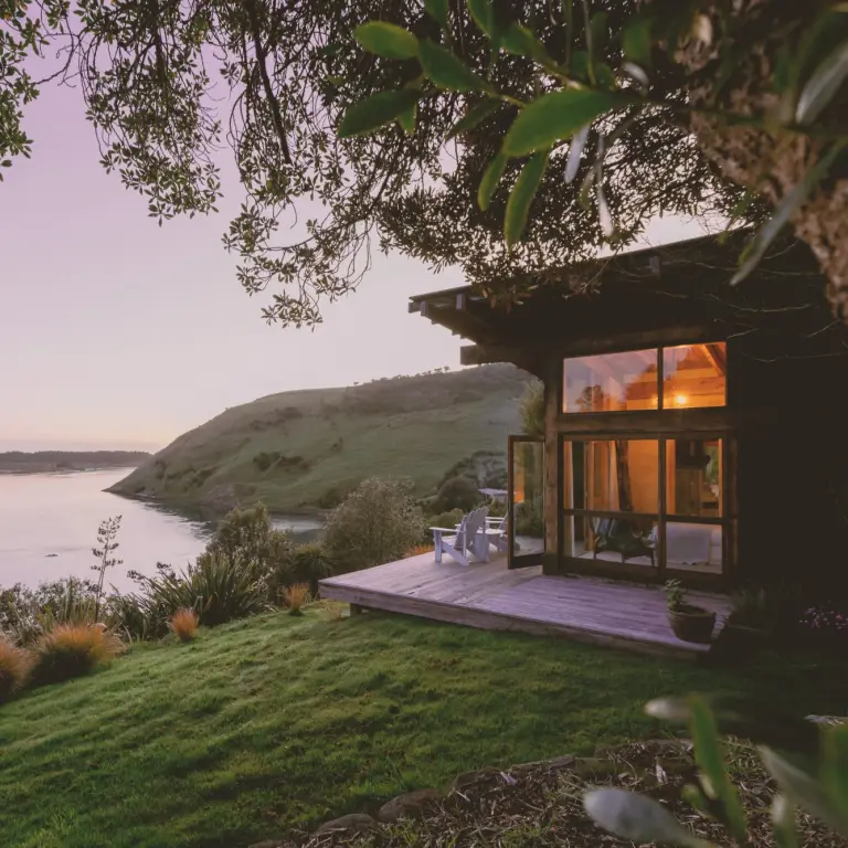 Ein abgeschiedenes Ferienhäuschen aus Holz mit einer Holzterrasse und zwei Stühlen, das an einem Hang thront und den Blick auf das ruhige Wasser der Otago-Halbinsel in Neuseeland bietet – in der Abenddämmerung, mit warm leuchtenden Innenräumen