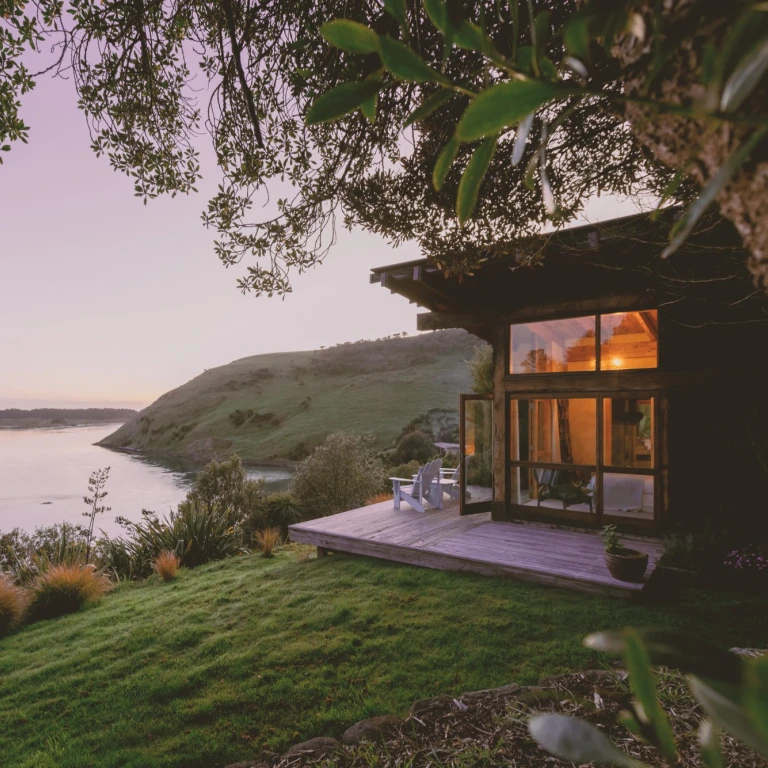 Ein abgeschiedenes Ferienhäuschen aus Holz mit einer Holzterrasse und zwei Stühlen, das an einem Hang thront und den Blick auf das ruhige Wasser der Otago-Halbinsel in Neuseeland bietet – in der Abenddämmerung, mit warm leuchtenden Innenräumen