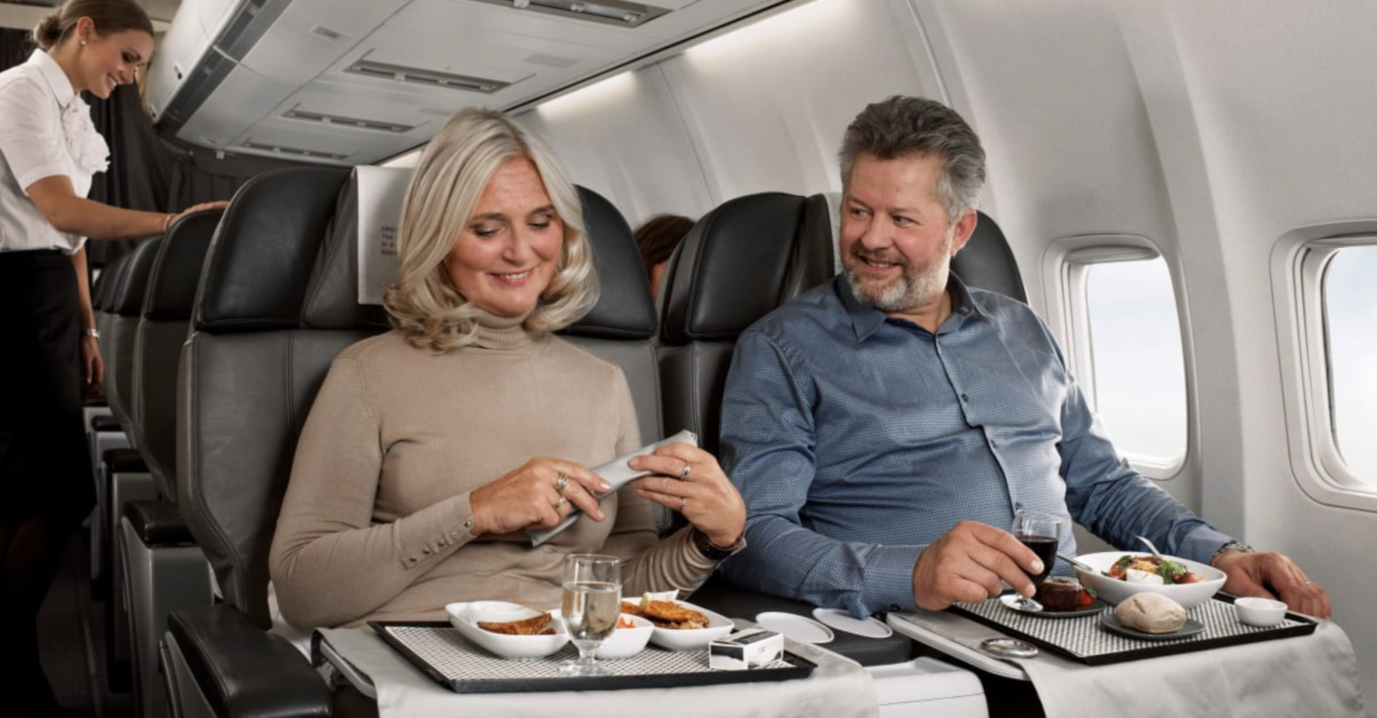Couple enjoying a meal in premium economy seats on a long-haul flight, with a flight attendant serving in the background