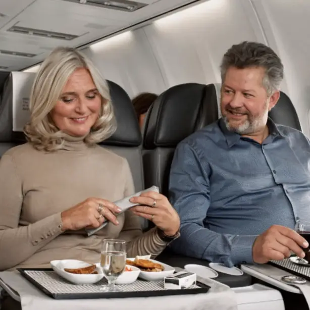 Couple enjoying a meal in premium economy seats on a long-haul flight, with a flight attendant serving in the background