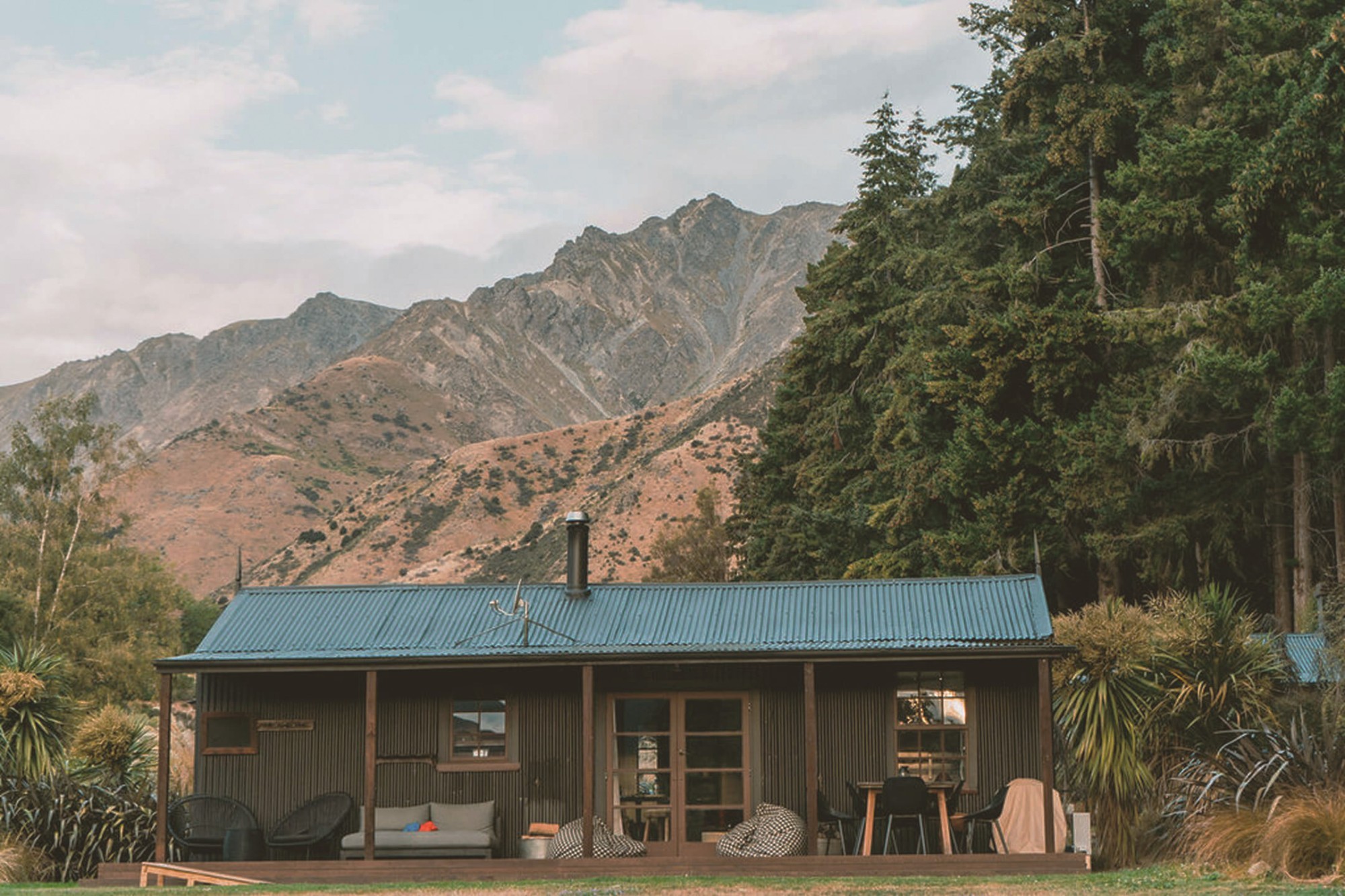 Rustikales Ferienhaus aus Wellblech mit überdachter Veranda und Sitzgelegenheiten im Freien, vor der Kulisse dramatischer Felsgipfel in der Nähe des Lake Hawea, Wanaka, Neuseeland