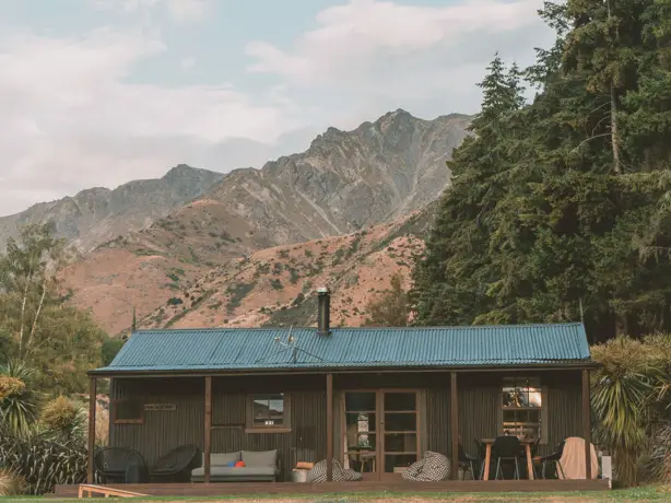 Rustikales Ferienhaus aus Wellblech mit überdachter Veranda und Sitzgelegenheiten im Freien, vor der Kulisse dramatischer Felsgipfel in der Nähe des Lake Hawea, Wanaka, Neuseeland