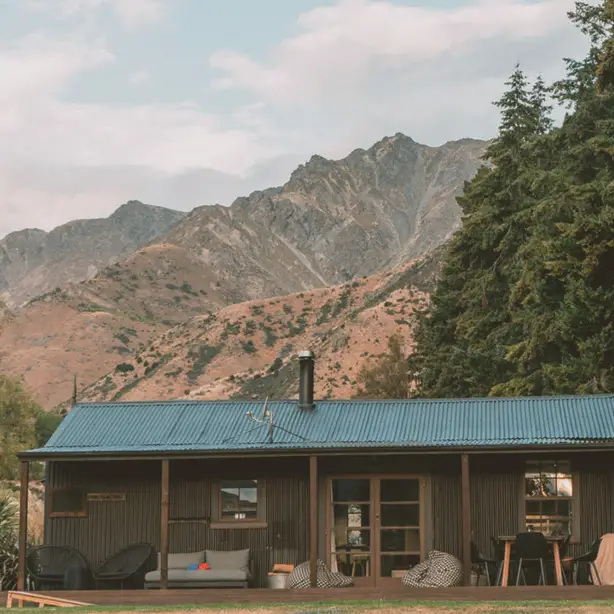 Rustic corrugated iron farmstay cottage with covered veranda and outdoor seating, set against dramatic rocky mountain peaks near Lake Hawea, Wanaka, New Zealand
