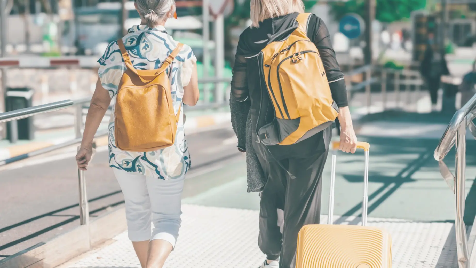 Two people walking to an airport wearing backpacks and one has a pull along suitcase