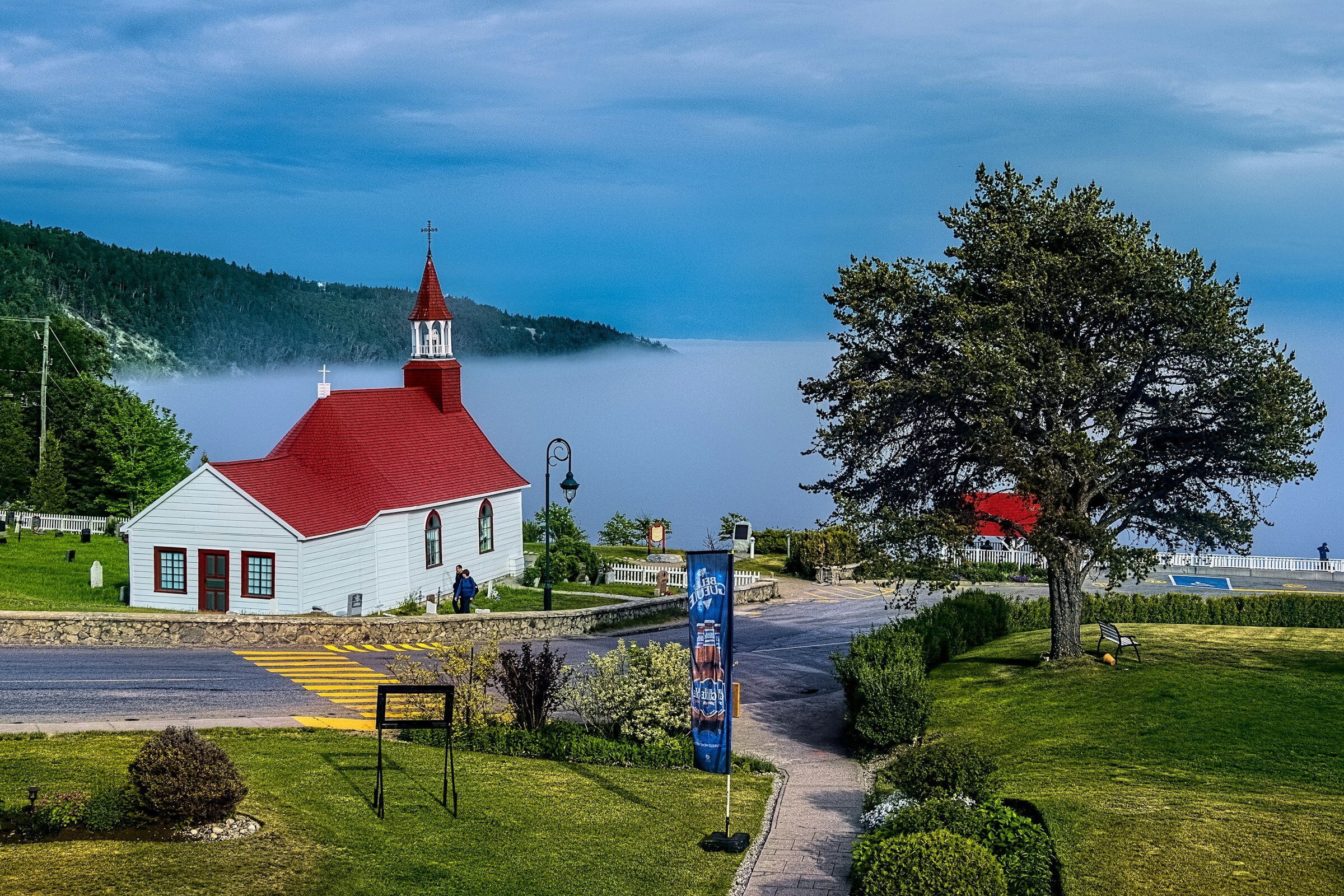 A small white church with a red roof overlooking a misty bay in Canada, surrounded by green lawns and forested hills.