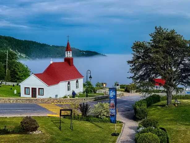 Eine kleine weiße Kirche mit rotem Dach mit Blick auf eine neblige Bucht in Kanada, umgeben von grünen Wiesen und bewaldeten Hügeln.