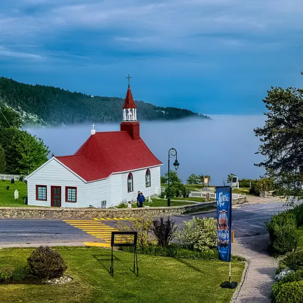 A small white church with a red roof overlooking a misty bay in Canada, surrounded by green lawns and forested hills.