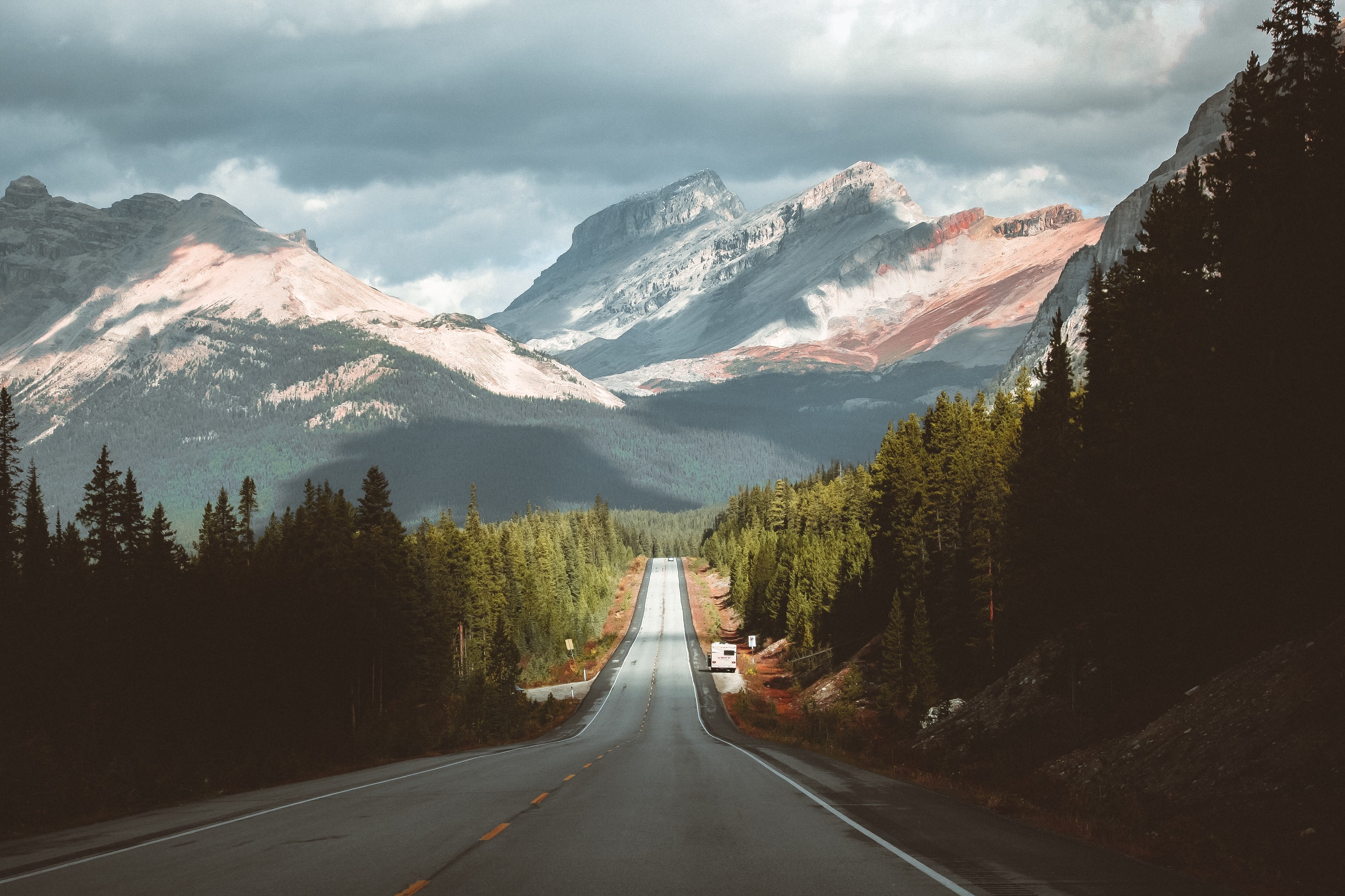 Der Icefields Parkway mit Bäumen zu beiden Seiten der Straße und schneebedeckten Bergen in der Ferne, unter einem stimmungsvollen Himmel