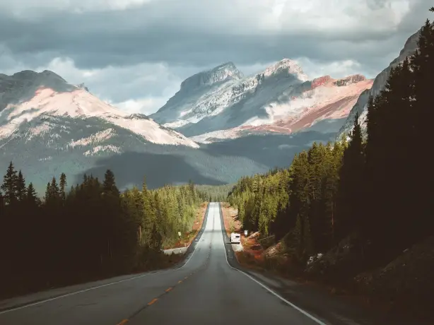 Der Icefields Parkway mit Bäumen zu beiden Seiten der Straße und schneebedeckten Bergen in der Ferne, unter einem stimmungsvollen Himmel