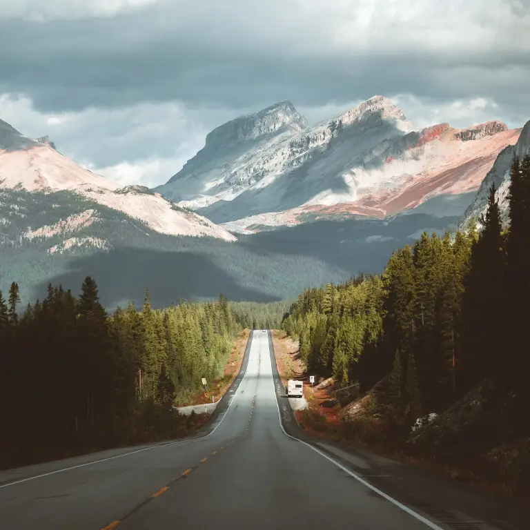 Der Icefields Parkway mit Bäumen zu beiden Seiten der Straße und schneebedeckten Bergen in der Ferne, unter einem stimmungsvollen Himmel