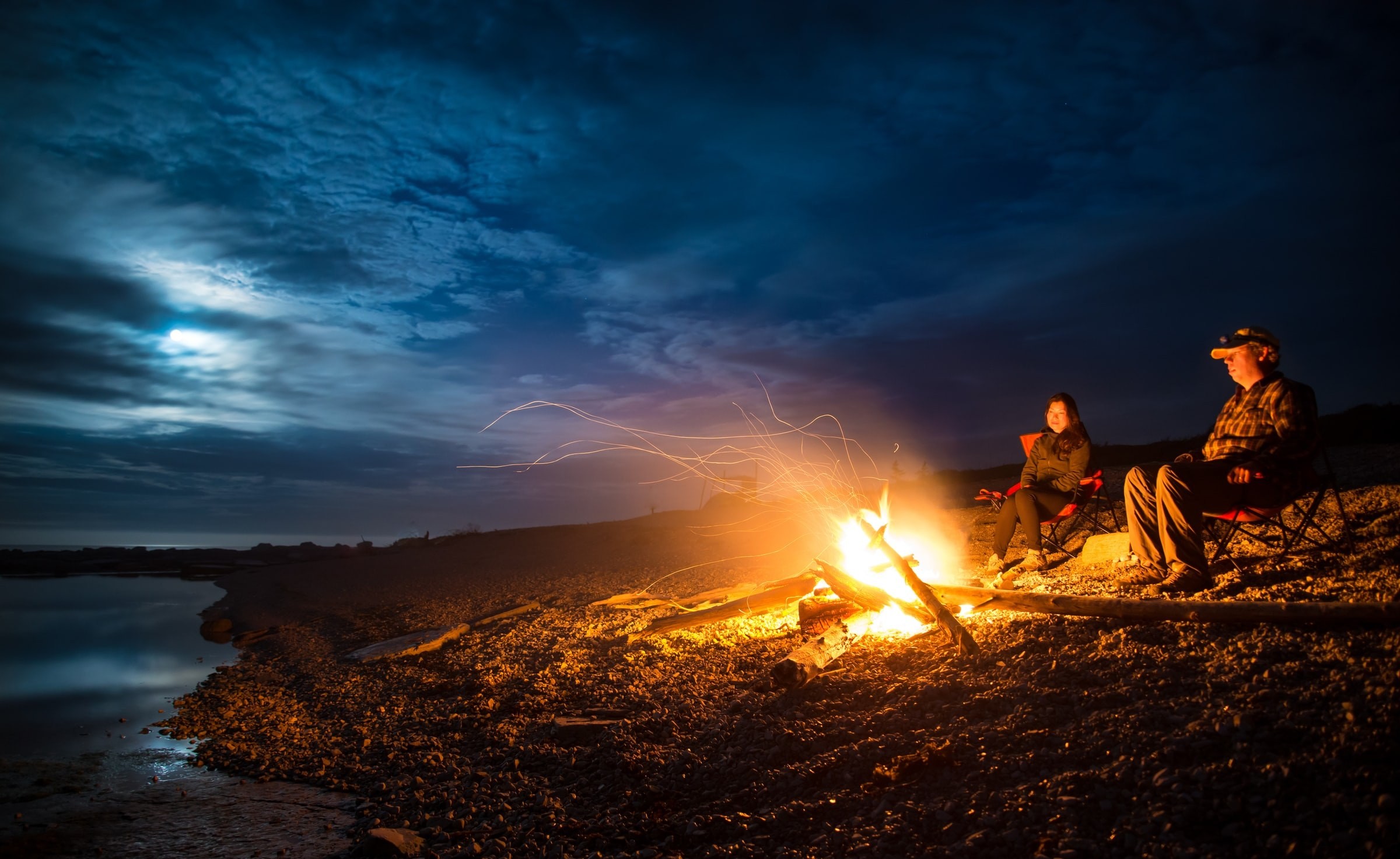 Ein Mann und eine Frau sitzen am Lagerfeuer an einem Gewässer in der Dunkelheit