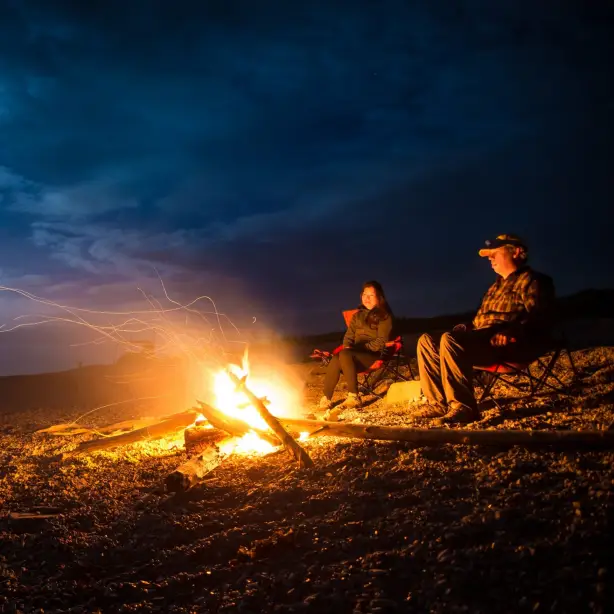 Ein Mann und eine Frau sitzen am Lagerfeuer an einem Gewässer in der Dunkelheit