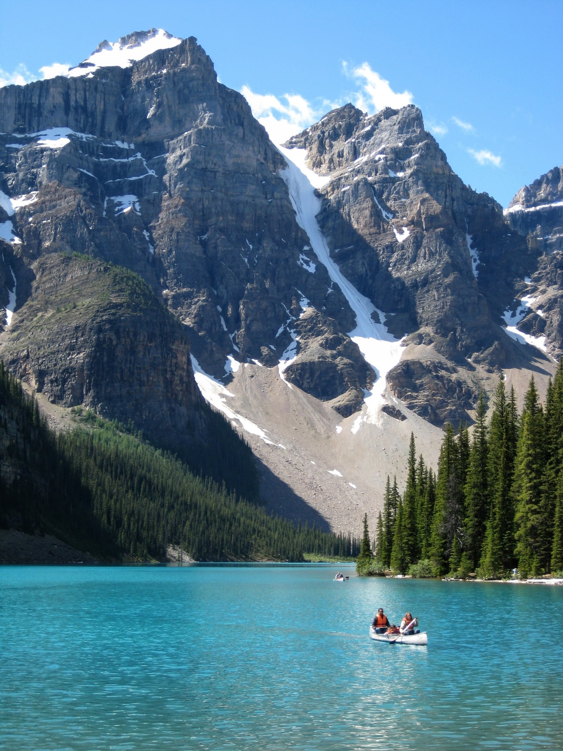 Zwei Personen paddeln im Kanu auf dem leuchtend türkisfarbenen Wasser des Moraine Lake, umgeben von hohen Nadelbäumen und dramatischen schneebedeckten Felsgipfeln des Valley of the Ten Peaks, im Banff-Nationalpark, Alberta, Kanada.