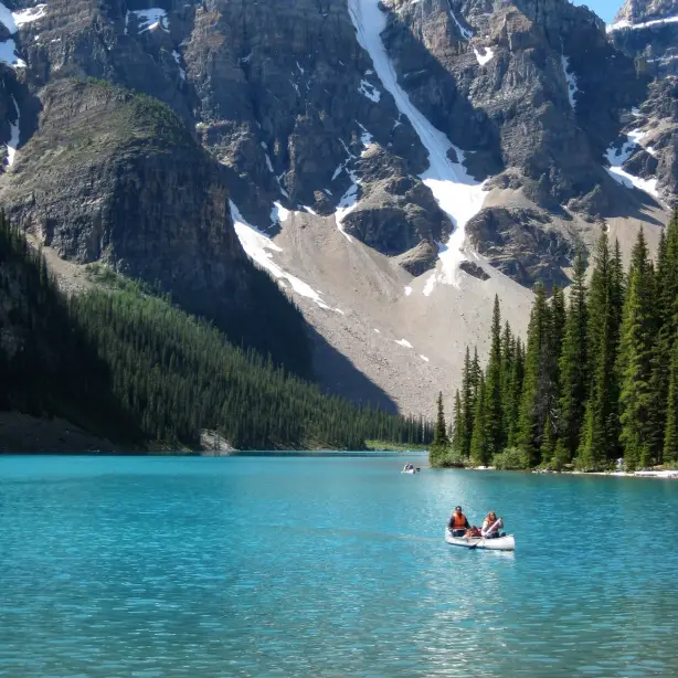 Zwei Personen paddeln im Kanu auf dem leuchtend türkisfarbenen Wasser des Moraine Lake, umgeben von hohen Nadelbäumen und dramatischen schneebedeckten Felsgipfeln des Valley of the Ten Peaks, im Banff-Nationalpark, Alberta, Kanada.