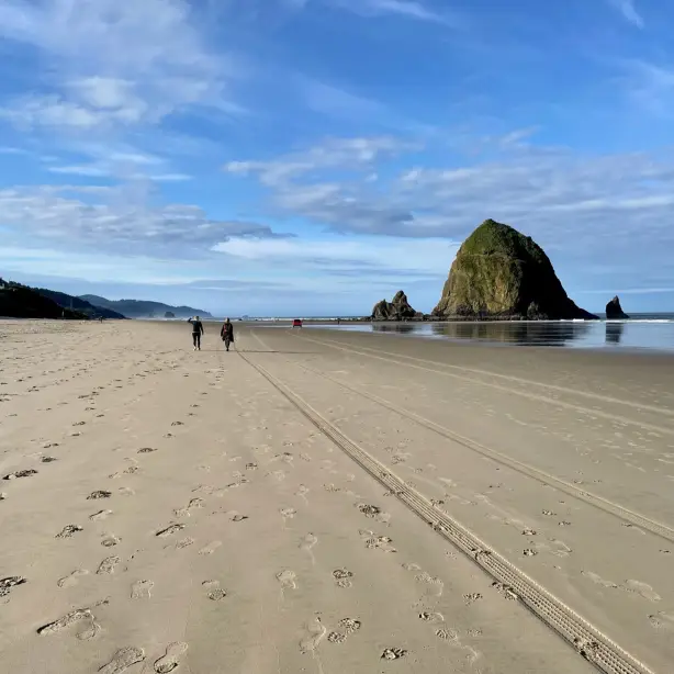 Twee mensen lopen over het strand van Cannon Beach richting een grote rotsformatie die uit de zee oprijst.