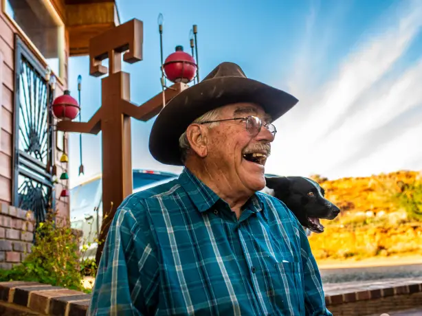 Der pensionierte Cowboy und Besitzer einer Gäste-Ranch Jerry Kinknade schaut nach rechts von der Kamera und lächelt, sein Hund und seine Ranch sind hinter ihm zu sehen.