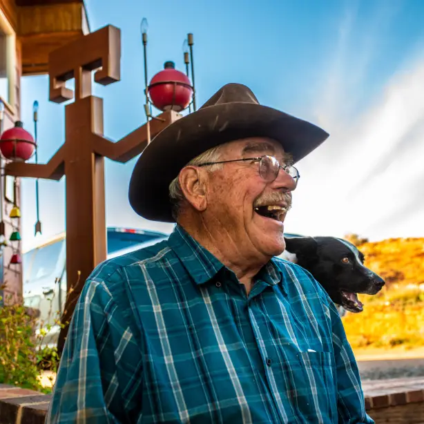 Retired cowboy and guest ranch owner Jerry Kinknade looks to the right of the camera and smiles, his dog and ranch are behind him