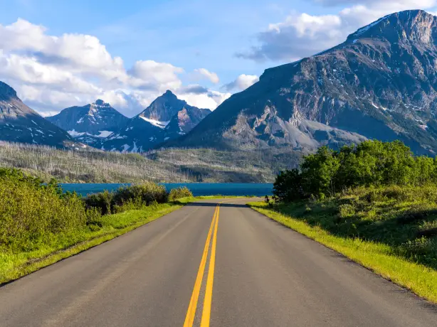Eine offene Straße, die zu den Bergen und dem See im Glacier National Park, Montana, USA, führt, unter einem strahlenden Sommerhimmel.