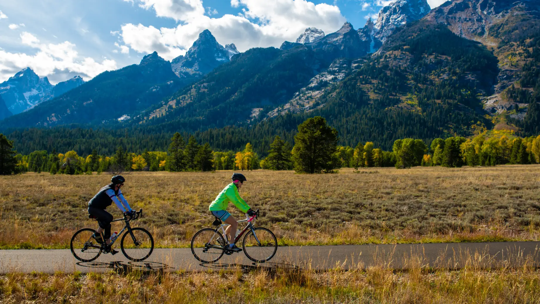 Zwei Menschen radeln auf einem Weg vor dem Hintergrund üppig grüner und schneebedeckter Berge im Grand-Teton-Nationalpark, Wyoming, USA.