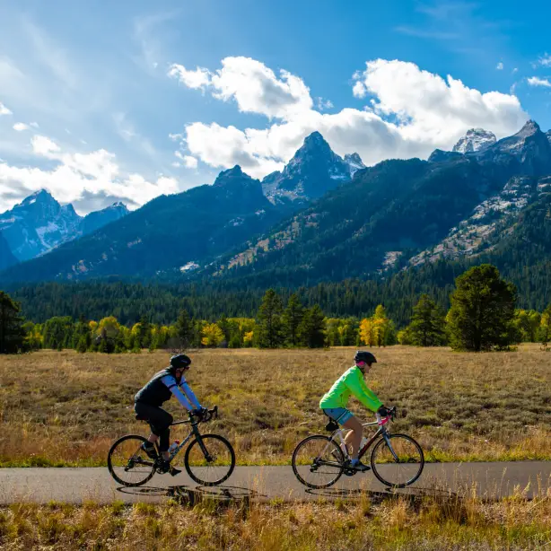 Zwei Menschen radeln auf einem Weg vor dem Hintergrund üppig grüner und schneebedeckter Berge im Grand-Teton-Nationalpark, Wyoming, USA.
