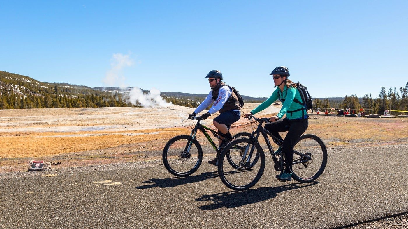 Zwei Menschen fahren mit dem Fahrrad an einem dampfenden Geysir im Yellowstone-Nationalpark in den USA vorbei
