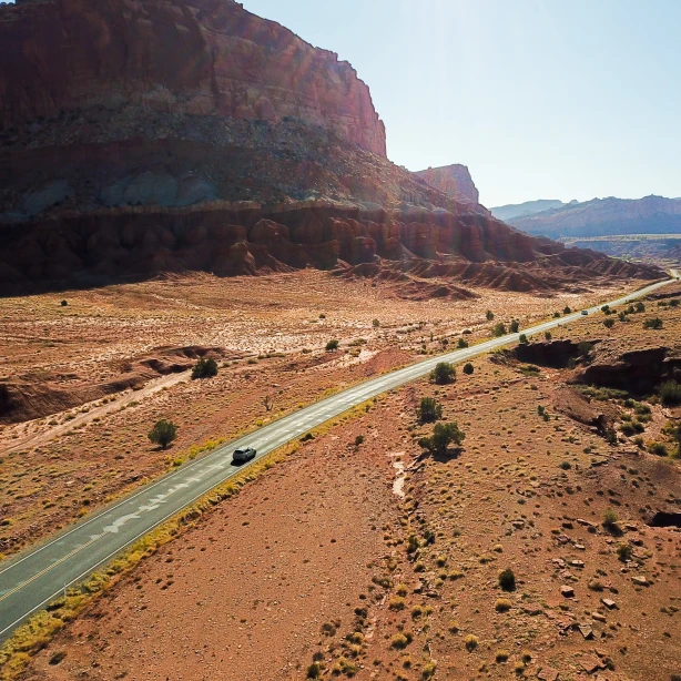 Ein Auto fährt auf einem abgelegenen Wüsten-Highway in Utah, umgeben von roten Felsen und weiten Landschaften.