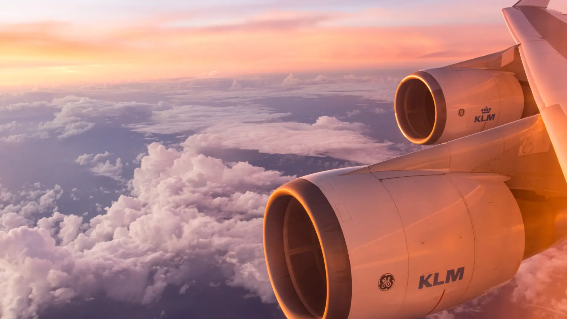 Flügel und Triebwerke eines KLM-Flugzeugs leuchten im weichen Licht eines Sonnenaufgangs über einer Wolkenschicht.