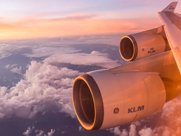 Flügel und Triebwerke eines KLM-Flugzeugs leuchten im weichen Licht eines Sonnenaufgangs über einer Wolkenschicht.