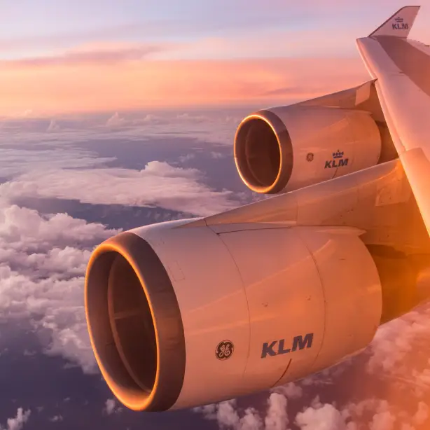 Flügel und Triebwerke eines KLM-Flugzeugs leuchten im weichen Licht eines Sonnenaufgangs über einer Wolkenschicht.