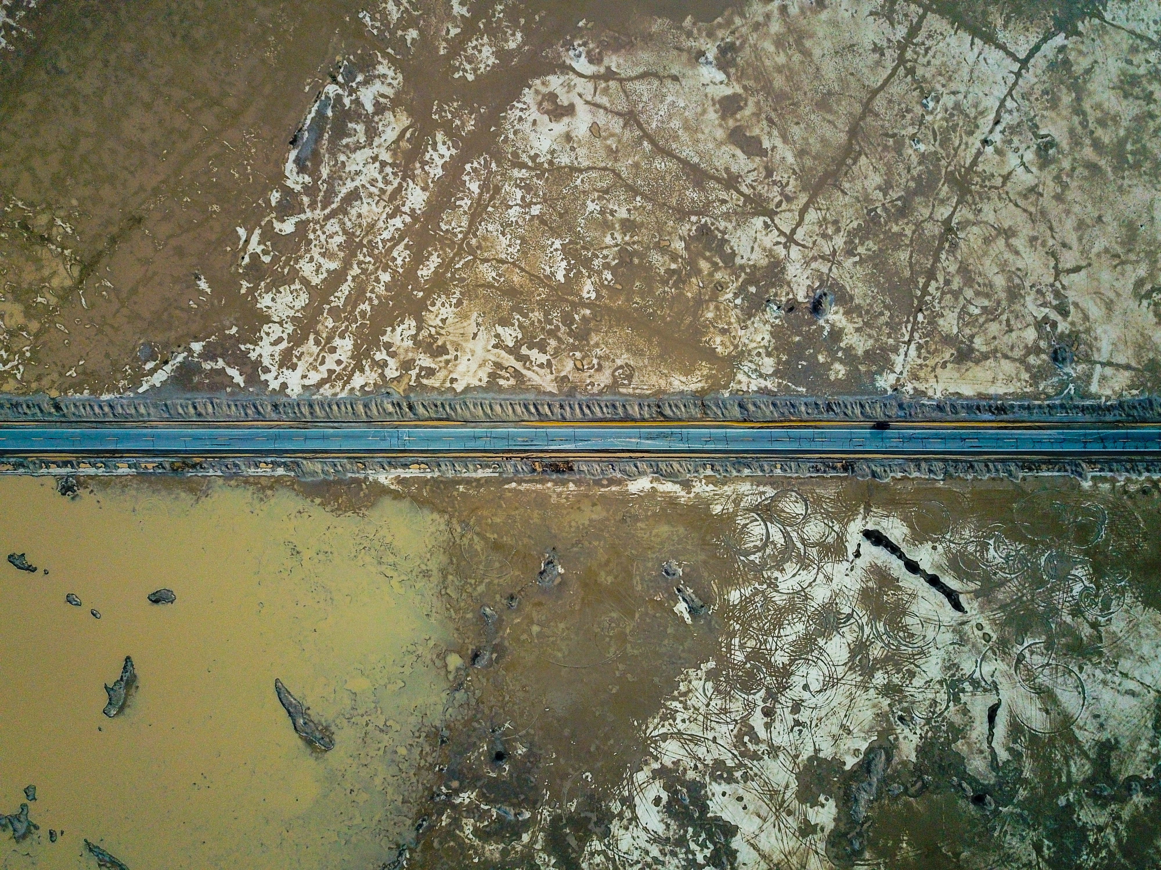 Eine gerade Straße, die durch die kargen Wüstenebenen des Death Valley führt, von oben gesehen, mit Wasserflecken und strukturierter, rissiger Erde auf beiden Seiten.