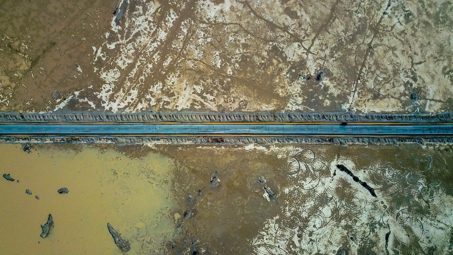 A straight road cutting through the barren desert flats of Death Valley, seen from above with patches of water and textured, cracked earth on either side.