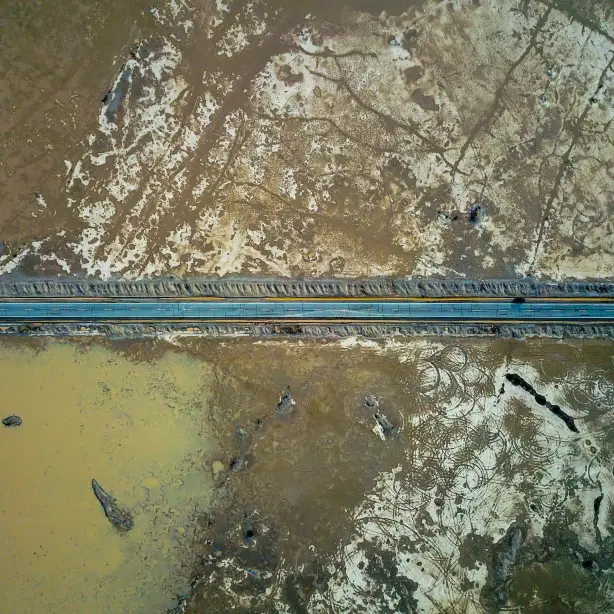 Een rechte weg dwars door de dorre woestijnvlakte van Death Valley, van bovenaf gezien met waterpartijen en gebarsten aarde aan weerszijden.