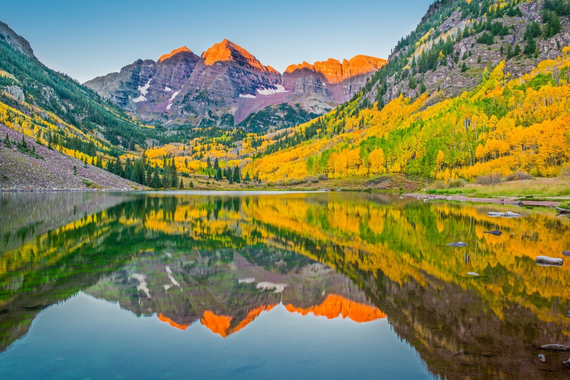 Von der Sonne gerötetes Bergpanorma und herbstlich bunte Bäume mit Spiegelung in einem See
