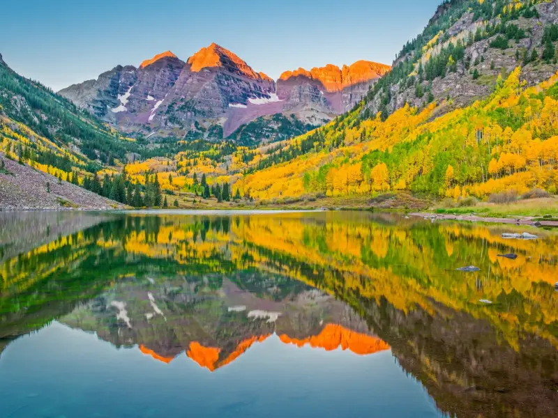 Von der Sonne gerötetes Bergpanorma und herbstlich bunte Bäume mit Spiegelung in einem See