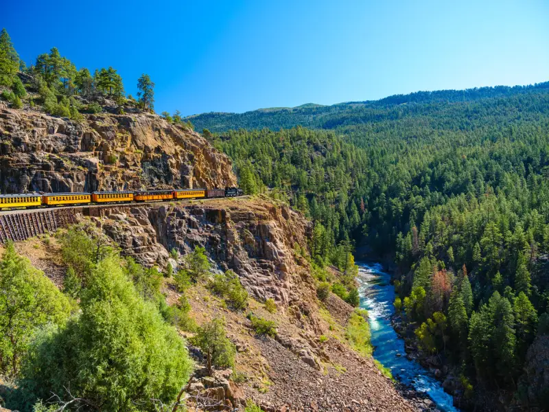 Historischer Zug fährt auf einer schmalen Trasse entlang steiler Felsklippen durch eine bewaldete Berglandschaft in Colorado, während tief unten ein türkisblauer Fluss durch die Schlucht fließt.