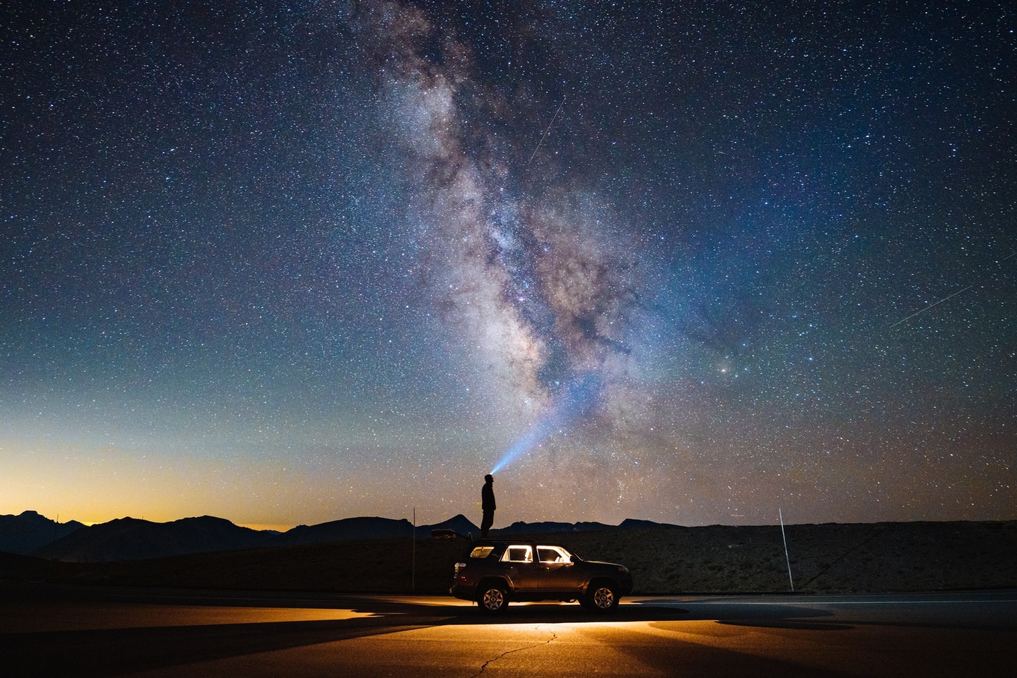 Eine Person steht nachts auf dem Dach eines Geländewagens in der Berglandschaft von Colorado und richtet eine Taschenlampe in den sternenklaren Himmel. Über ihr erstreckt sich die deutlich sichtbare Milchstraße, während die Silhouetten der Berge am Horizont zu erkennen sind.