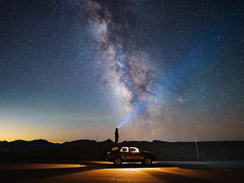 Eine Person steht nachts auf dem Dach eines Geländewagens in der Berglandschaft von Colorado und richtet eine Taschenlampe in den sternenklaren Himmel. Über ihr erstreckt sich die deutlich sichtbare Milchstraße, während die Silhouetten der Berge am Horizont zu erkennen sind.