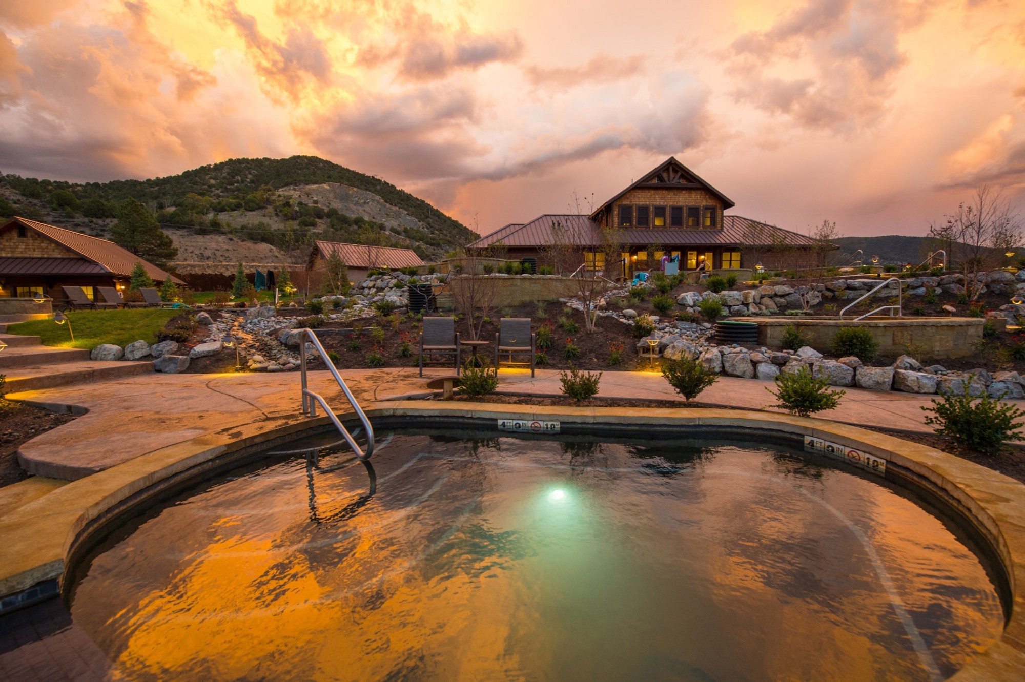 Thermalbecken der Iron Mountain Hot Springs in Glenwood Springs, Colorado, bei Sonnenuntergang. Im Vordergrund spiegelt sich der farbenfrohe Himmel im warmen Wasser, dahinter liegen das Spa-Gebäude, gepflegte Gartenanlagen und die umliegenden Hügel.